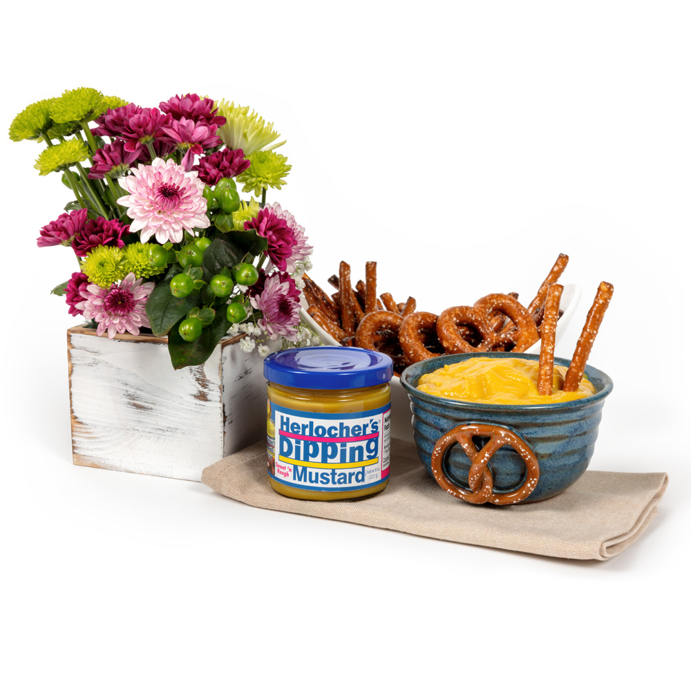 Blue handcrafted pottery crock filled with mustard and pretzel sticks, served with hard pretzels and a jar of Herlocher’s dipping mustard beside a rustic floral arrangement with pink, purple, and green flowers on a light background.